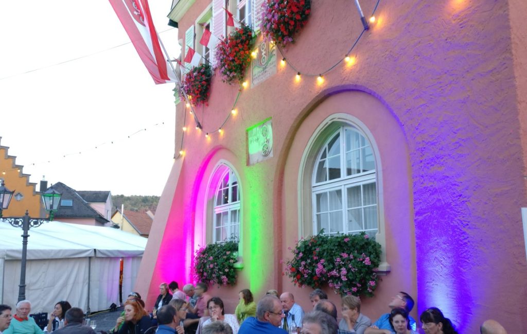 Das Rathaus ist beleuchtet. Am Fuße seiner Mauern sitzt der Rheinhesse und erfreut sich am Gespräch mit dem Tischnachbarn und dem Wein.